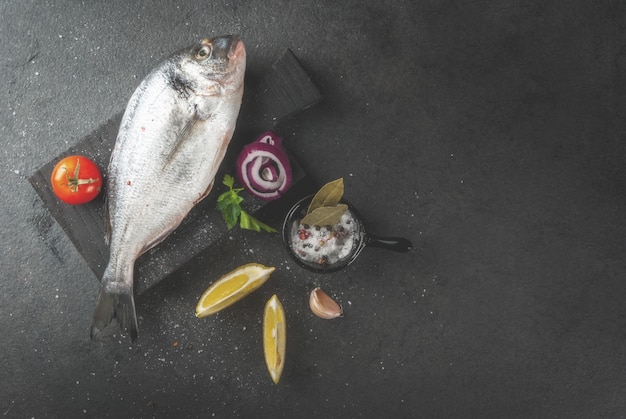 Peixe orgânico cru fresco dorado, pargo com ingredientes para cozinhar limão, tomate, cebola, sal, pimenta, folha de louro, verduras, óleo. na tábua de madeira velha, na mesa de pedra preta