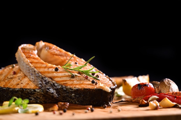 Foto peixe salmão grelhado e vários legumes na mesa de madeira em preto