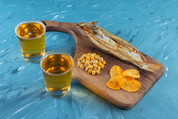 Peixe seco, batatas fritas, grão de bico em uma tábua ao lado de um copo de cerveja, na superfície azul.