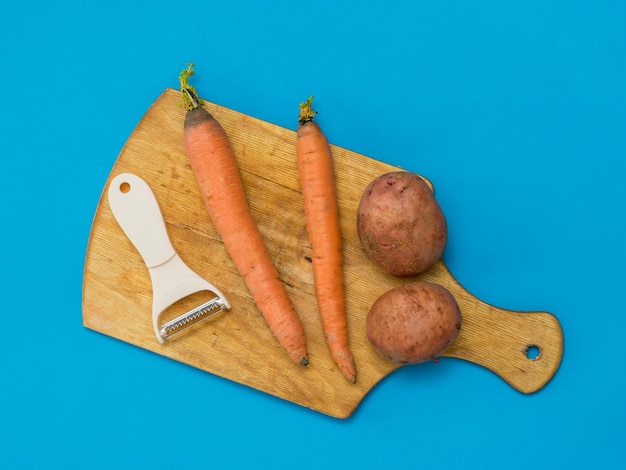 Pelador de verduras, patatas y zanahorias sobre una tabla de cortar sobre un fondo azul. Quipment especial para verduras magras.