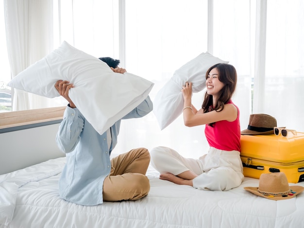 Pelea de almohadas de pareja Mujer y hombre asiáticos hermosos con camisa de mezclilla sentados y peleando juntos con almohadas cerca de la maleta amarilla en la cama en la luminosa habitación del hotel Felices vacaciones