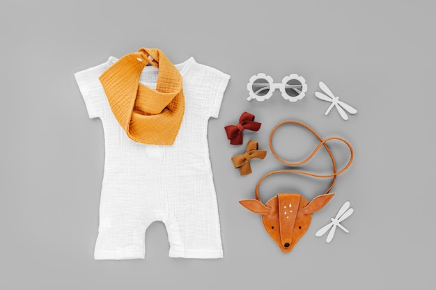 Pelele blanco, babero naranja con bolso infantil y gafas de sol. Conjunto de ropa de bebé y accesorios para vacaciones de verano sobre fondo gris. Traje de moda infantil. Endecha plana, vista superior