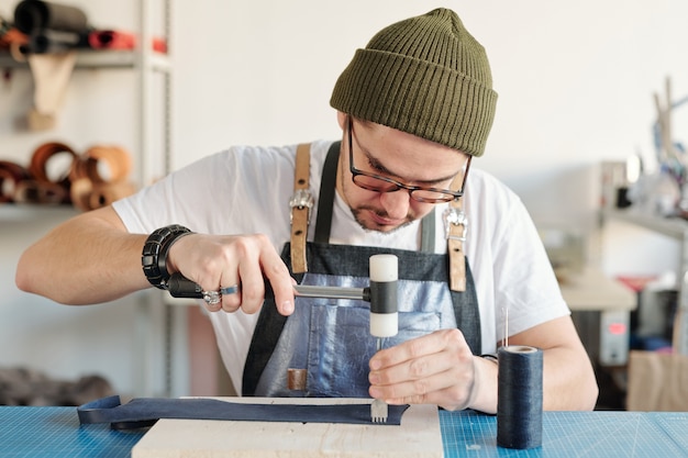 Foto peletero contemporáneo o maestro de la artesanía utilizando herramientas manuales para procesar la pieza de cuero negro sobre una tabla de madera