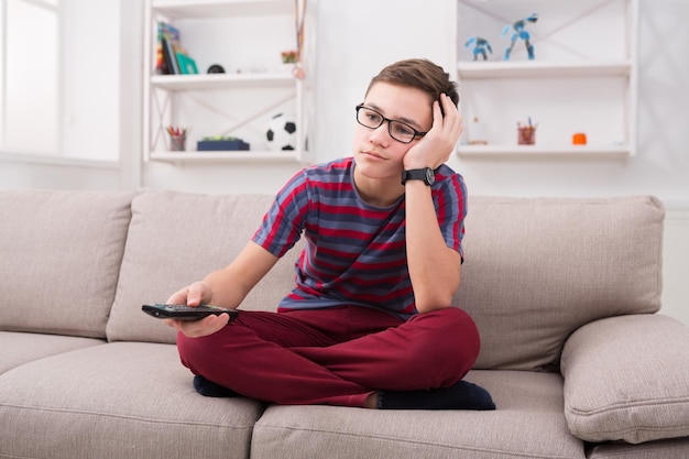 Película aburrida. Adolescente sosteniendo el control remoto y mirando aburrido mientras está sentado en el sofá en casa en la sala de estar