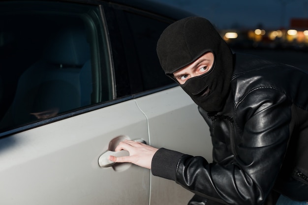 Peligro de robo de automóviles, concepto de publicidad de seguros de automóviles. Ladrón masculino con pasamontañas en la cabeza intentando abrir la puerta del coche. Vehículo de desbloqueo de carjacker