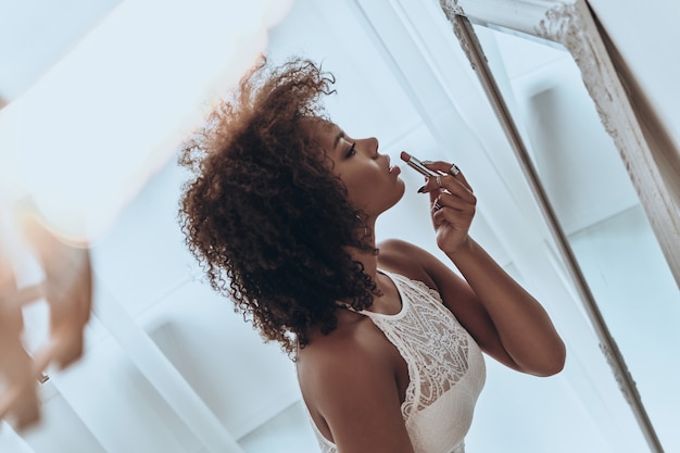 Peligrosamente hermosa. Vista superior de la atractiva joven africana poniéndose lápiz labial mientras está de pie frente al espejo en casa