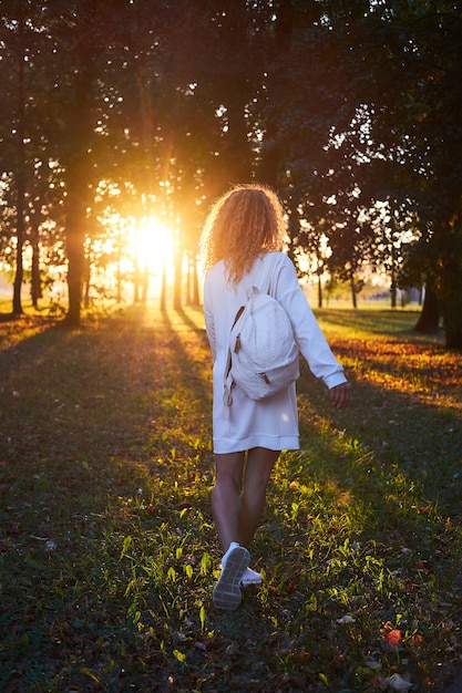 Pelirroja jengibre rizada, vestida con ropa blanca y mochila blanca, diviértete a la luz del atardecer