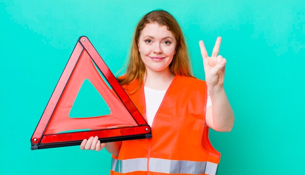 Foto pelirroja mujer bonita sonriendo y luciendo amigable mostrando el cocnept de accidente de triángulo de coche número dos