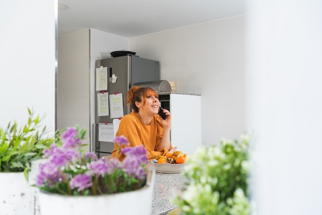 Pelirroja mujer caucásica en una camisa naranja hablando por teléfono en una cocina