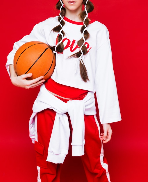 Pelota de baloncesto en manos de una niña vestida con ropa deportiva. Foto de estudio sobre fondo rojo. Foto vertical