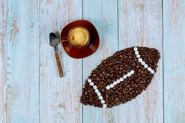 Pelota de fútbol americano hecha de granos de café con una taza de café recién hecho en la superficie de madera azul.