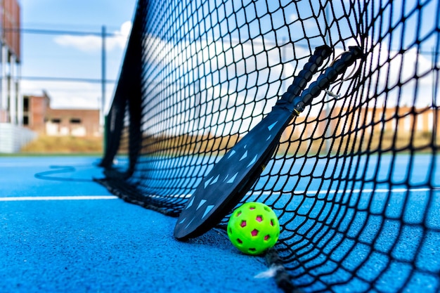 Foto pelota de pickleball y paletas descansando sobre la red de una cancha de pickleball