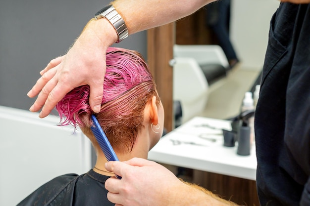 Una peluquera está peinando el cabello corto húmedo teñido de rosa de la clienta en la vista posterior de la peluquería