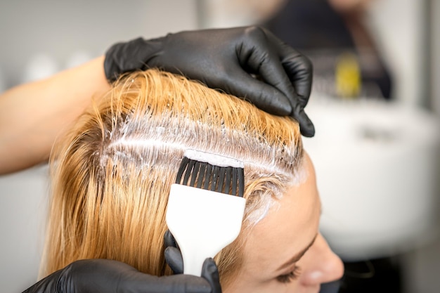 La peluquera está tiñendo las raíces del cabello rubio con un cepillo para una mujer joven en una peluquería