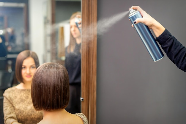 Una peluquera está usando spray para el cabello para arreglar el peinado corto de la joven morena sentada en la peluquería