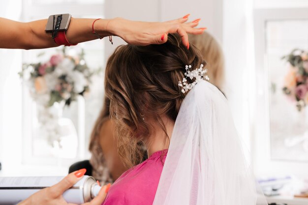 Una peluquera hace un peinado elegante para peinar a una novia con un velo blanco en el pelo en el salón