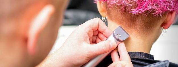 Peluquería afeitado nuca y cuello con recortadora eléctrica de una joven mujer caucásica con pelo corto de color rosa en un salón de belleza.