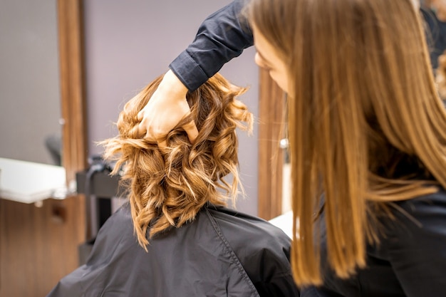 Peluquería femenina comprueba el peinado rizado marrón de una joven mujer caucásica en un salón de belleza