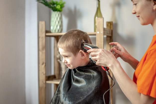 Peluquería joven moderna está cortando el cabello de un niño pequeño con una cortadora de cabello