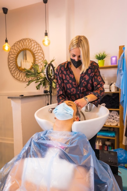 Peluquería con máscara de protección facial lavando el cabello del cliente con agua y jabón Apertura con medidas de seguridad de Peluquerías en la pandemia de Covid19