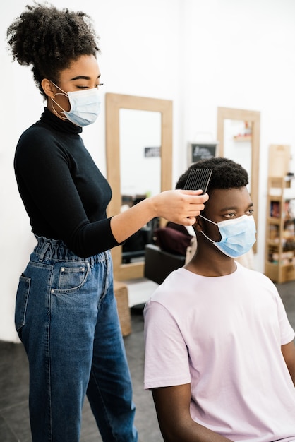 Peluquero afroamericano de belleza peinando y peinando a un cliente afroamericano con máscara y guantes para protegerse de la pandemia de coronavirus