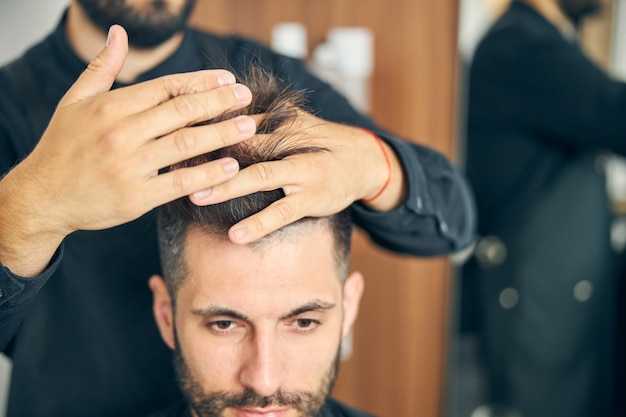 Peluquero barbudo profesional de pie detrás de su cliente y comprobando la longitud del cabello