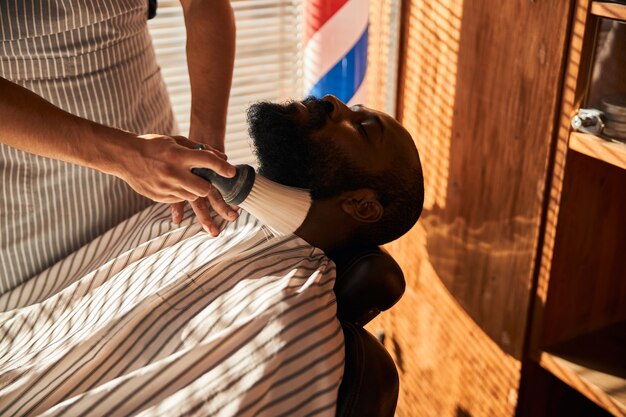 Peluquero cepillando el cabello alrededor del escote del cliente después de recortar la barba