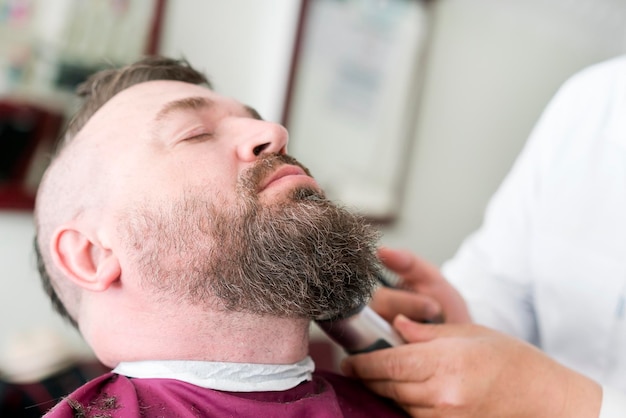 Un peluquero corta la barba con un cortapelos de un hombre adulto