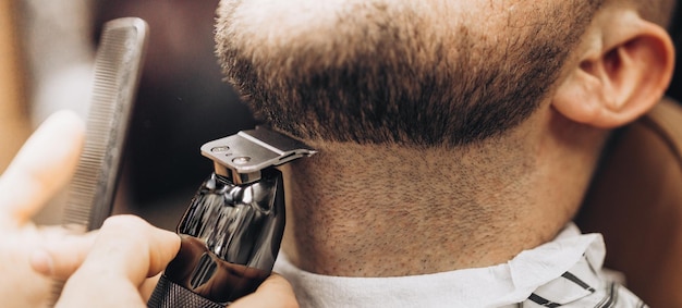 Peluquero cortando barba al hombre en la barbería enmarcando la maquinilla de afeitar eléctrica de la línea del cabello marrón teñido vintage