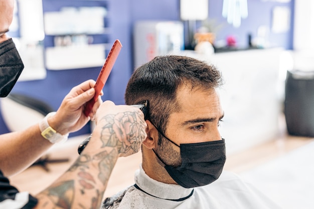 Foto peluquero cortando el cabello de un cliente caucásico con una máscara con una máquina en una barbería