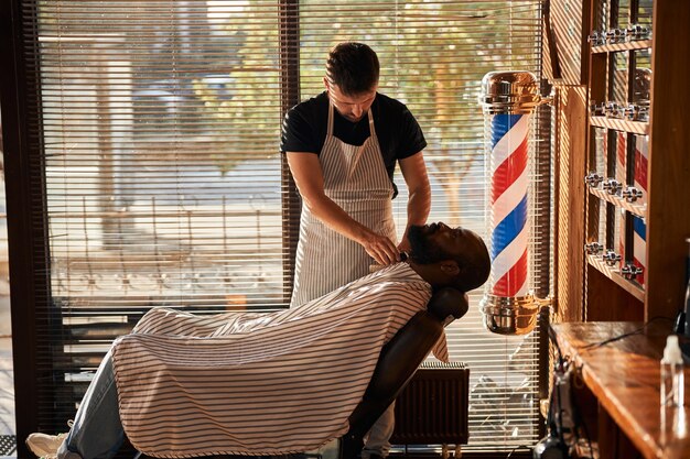 Peluquero en delantal recortando la barba del cliente en peluquería