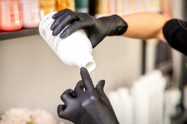 Un peluquero con guantes negros está preparando tinte para el cabello con una botella en una peluquería de cerca