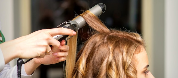 El peluquero hace rizos con un rizador para la joven con cabello castaño largo en un salón de belleza.