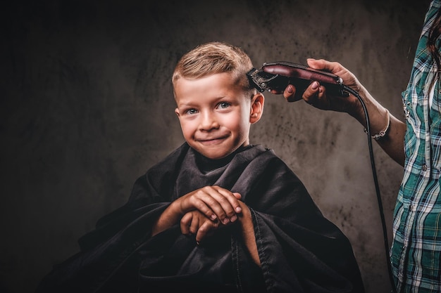 El peluquero infantil con el cortapelos está cortando a un niño pequeño contra un fondo oscuro. Lindo niño preescolar cortándose el pelo.