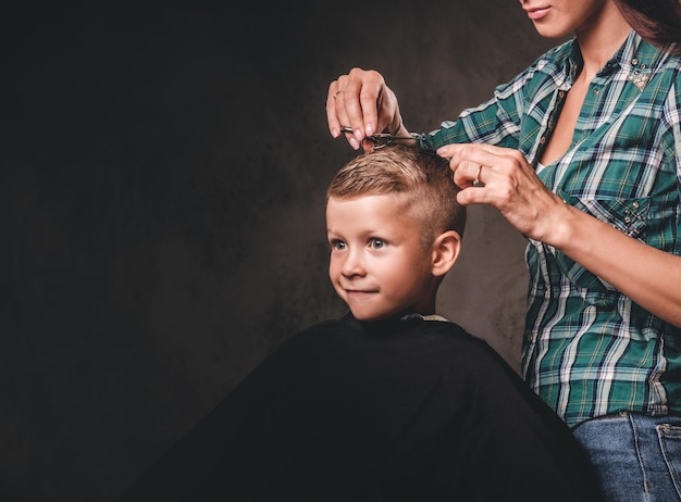 El peluquero infantil con tijeras está cortando a un niño pequeño contra un fondo oscuro. Un niño preescolar lindo y contento cortándose el pelo.