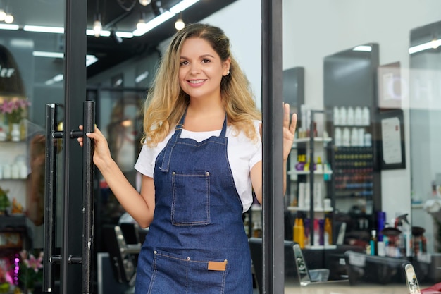 Peluquero joven positivo en delantal de mezclilla abriendo la puerta del salón para dejar entrar a los clientes