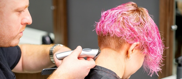El peluquero masculino afeita el cuello de una joven mujer caucásica con un peinado rosa corto con una afeitadora eléctrica en una peluquería de cerca