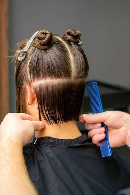 Foto peluquero masculino cortando el cabello de mujer joven con peine en peluquería. vista trasera