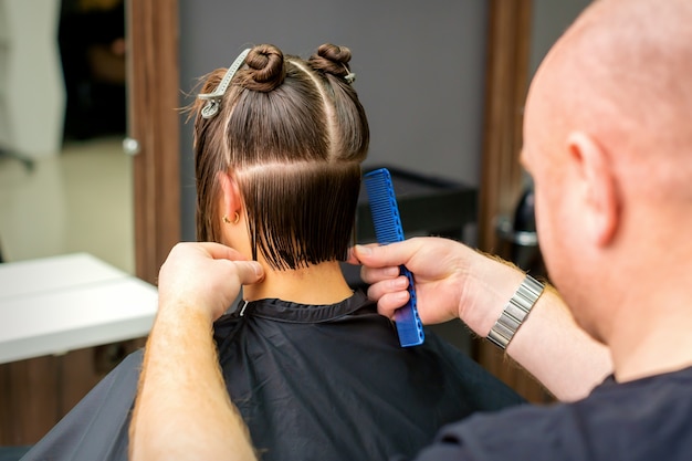 Peluquero masculino cortando el cabello de mujer joven con peine en peluquería.