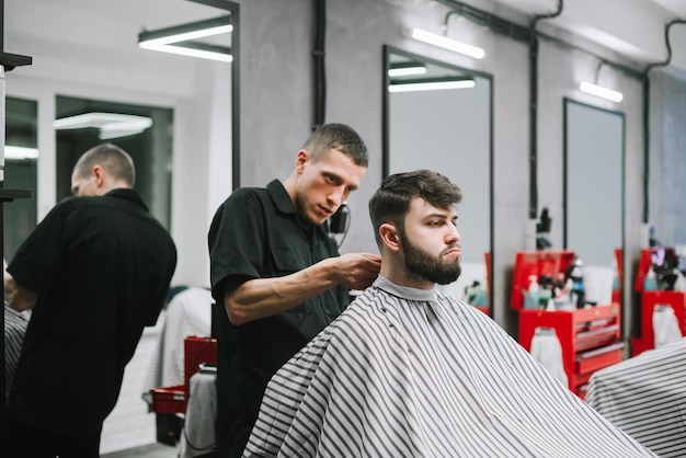 peluquero masculino crea un peinado elegante para un cliente barbudo descontento en una peluquería ligera