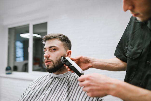 Peluquero profesional hace un corte de pelo de barba para un cliente con un cortapelos