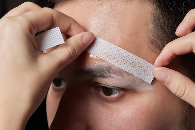 Peluquero profesional haciendo procedimiento de enhebrado y corrigiendo la forma de las cejas a un joven cliente masculino con pinzas en la peluquería Peluquería en el trabajo Concepto de cuidado de cejas Depilación de cejas