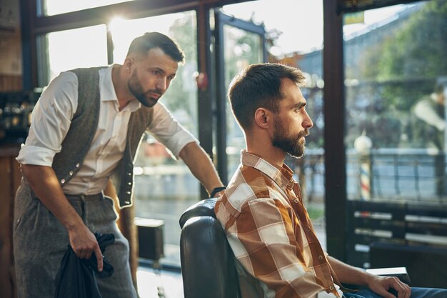 Peluquero profesional inspeccionando cuidadosamente el cabello de su cliente mientras está de pie detrás de su sillón