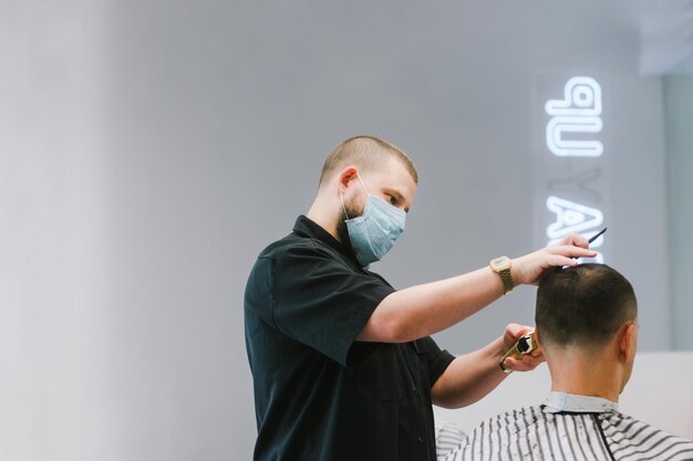 Peluquero profesional con una máscara de gasa médica en la cara haciendo un peinado para un cliente durante la cuarentena Trabajo de la barbería en el coronavirus el uso de equipo de protección personal