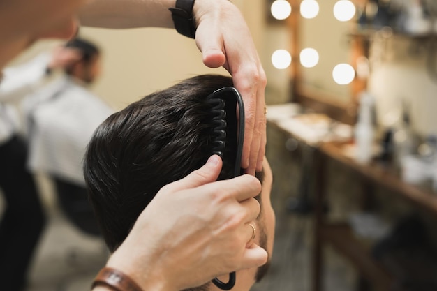 Peluquero profesional masculino hace peinado a hombre barbudo en peluquería, vista posterior, mano del peluquero con maquinilla.