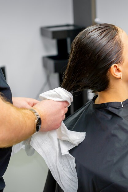 El peluquero seca el cabello lavado con una toalla a una hermosa joven morena en un salón de belleza