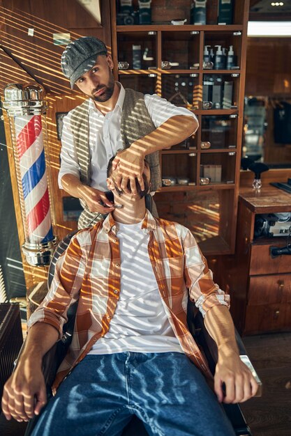 Peluquero serio sosteniendo una navaja de afeitar en el cuello de su cliente mientras lo afeita y mira a la cámara