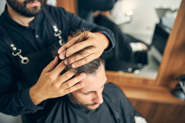 Peluquero serio vistiendo uniforme mientras trabaja en un salón moderno con el cliente