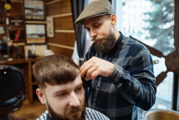 El peluquero sostiene el peine y corta el cabello del cliente. La peluquería profesional es una ocupación de moda. Peluquero masculino y cliente en salón de estilo retro