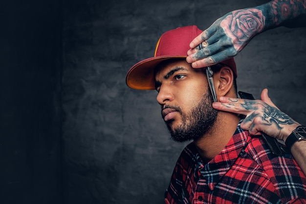 Peluquero tatuado cortando la barba de un hipster negro con una gorra de béisbol.